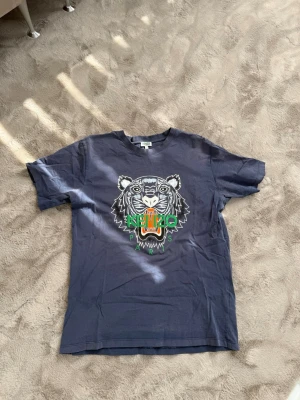 Mörkblå Kenzo t-shirt med tigertryck - Snygg mörkblå t-shirt från Kenzo med ikoniskt tigertryck framtill i vitt, grönt och orange. Klassisk passform och rund halsringning. Perfekt för dig som vill sticka ut med en cool streetstyle-look.