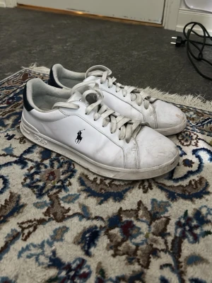 Vita Polo Ralph Lauren sneakers - Klassiska vita sneakers från Polo Ralph Lauren med lågt skaft och mörkblå detaljer vid hälen. Skorna har den ikoniska polospelaren på sidan och är tillverkade i slätt syntetmaterial med vita snören och sula. Endast smutsiga nu inte slitna.