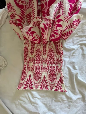 Vit och rosa kortklänning med volang - Snygg kortklänning i vitt och rosa med blommigt mönster och volang nertill. Klänningen har smockad överdel och är axelbandslös, perfekt för sommarens varma dagar. Materialet känns lätt och luftigt.