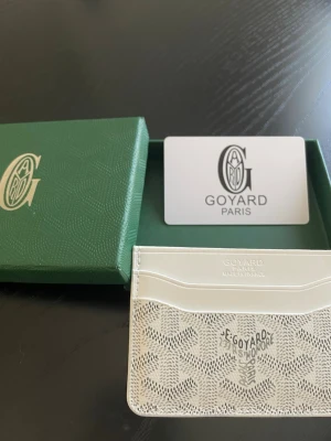 Vit kortfodral från Go4yard - Snyggt vitt kortfodral från Go4yard med klassiskt mönster och präglad logotyp framtill. Tillverkat i canvas och läder, har flera fack för kort och levereras med original grön ask. Perfekt för dig som vill ha en stilren och exklusiv accessoar.