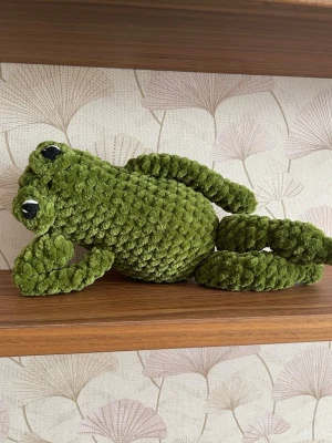 Virkad groda 28 cm handgjord - Handvirkad groda i mjukt chenillegarn med långa armar och ben samt broderade ögon, en lekfull och mysig figur som passar perfekt som present eller dekoration. Varje exemplar är unikt och gjort för hand. Höjd ca 28 cm. Mjuk, kramvänlig och lätt i vikt.