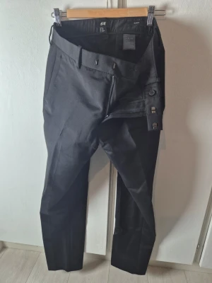 Svarta kostymbyxor från H&M Slim Fit - Snygga svarta kostymbyxor från H&M i slim fit-modell. Byxorna har pressveck, bälteshällor och stängs med dragkedja, hake och knapp. Materialet är en blandning av polyester och viskos som ger en mjuk och stilren känsla.