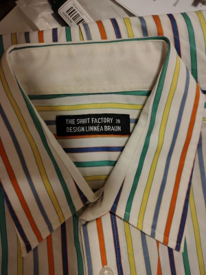 Randig skjorta från The Shirt Factory - Färgglad randig skjorta designad av Linnéa Braun för The Shirt Factory. Skjortan har vita, gröna, gula, blå och orange ränder samt klassisk krage och knappar framtill. Perfekt för dig som vill sticka ut lite extra.en special utgåva o limiterad upplaga