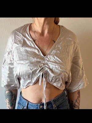 Beige croppad topp med snörning - Trendig croppad topp i beige med v-ringning och snörning framtill. Toppen har korta ärmar och är tillverkad i ett lätt, glansigt material som ger en cool look. Perfekt att matcha med höga jeans för en avslappnad stil.