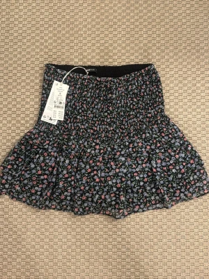 Blommig volangkjol från gina tricot  - Kort blommig kjol från gina tricot med volang och smockad midja. Kjolen har svart botten med små blommor i rosa, blått och grönt. Perfekt för sommarens alla tillfällen och har en luftig, lätt känsla.