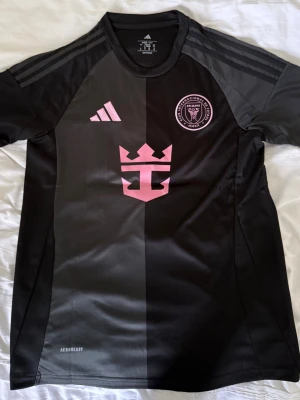 Inter Miami CF Messi matchtröja svart - Snygg svart matchtröja från Inter Miami CF med rosa detaljer, Messi och nummer 10 på ryggen. Tröjan har klubbmärke och Adidas-logga framtill samt AEROREADY-material som andas. Perfekt för fotbollsträning eller att bära som supporter.