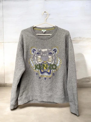 Grå Kenzo sweatshirt med tigertryck - Snygg grå sweatshirt från Kenzo med ikoniskt broderat tigerhuvud och texten 'KENZO PARIS' framtill. Tröjan har rund halsringning och långa ärmar. Perfekt för en trendig och avslappnad stil.
