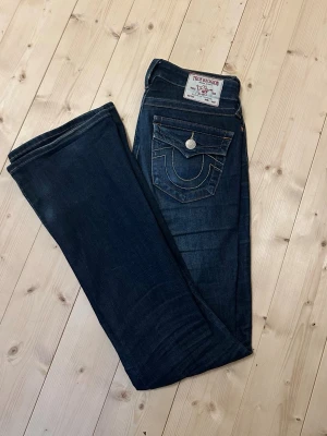 Mörkblå True Religion jeans - Säljer ett par mörkblå bootcut mid waist jeans  och klassiska fem fickor. Jeansen har snygga kontrastsömmar och stängs med dragkedja och knapp. Perfekta för en avslappnad och trendig look. Storlek W26 Becca bootcut. Använda men nästan inga tecken på användning, lite lite slitet längst ner på jeansen (se bild 7) Passar nån som är mellan 165-175. Säljer då de inte kommer till användning.
