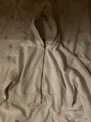 Vit croppad hoodie med dragkedja - Snygg vit hoodie med dragkedja framtill och stor huva. Modellen är croppad och har två stora fickor på magen. Perfekt för en chill och avslappnad stil. Materialet känns mjukt och bekvämt, troligen bomullsblandning.