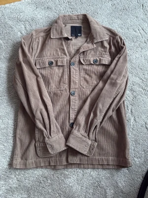 Beige manchester overshirt H&M - Säljer en beige overshirt i manchestertyg från H&M. Skjortan har två bröstfickor med knappar, klassisk krage och långa ärmar med manschetter. Perfekt lager på lager-plagg med avslappnad passform och snygga detaljer.