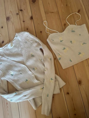 Beige ribbad kofta och linne med blommor - Säljer ett set med en beige ribbad kofta och matchande linne med tunna axelband. Båda plaggen har broderade gula rosor och gröna blad. Koftan har knappar framtill och lång ärm, perfekt nu för sommaren. Toppen är i storlek XS och koftan i storlek S! 