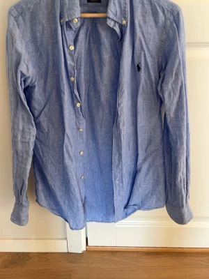 Blå linneskjorta från Ralph Lauren - Snygg blå skjorta i linne från Ralph Lauren med klassisk krage och knappar framtill. Skjortan har lång ärm och den ikoniska loggan broderad på bröstet. Perfekt för en stilren och avslappnad look.