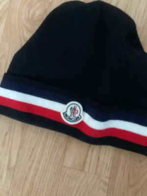 Svart Moncler mössa med ränder - Snygg svart mössa från Moncler med klassiska ränder i blått, vitt och rött runt kanten. Mössan har Moncler-logga framtill och en liten etikett på sidan. Perfekt accessoar för att hålla värmen och samtidigt se stilren ut.
