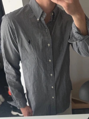 Svartvit rutig skjorta från Ralph Lauren - Klassisk svartvit rutig skjorta från Ralph Lauren med button-down krage och broderad logga på bröstet. Skjortan har lång ärm, normal passform och vita knappar framtill. Perfekt till jeans eller chinos för en clean och stilren look.