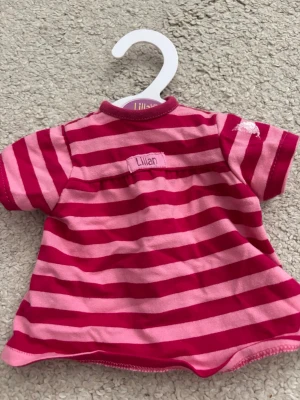 Randig dockklänning från Lillan - Söt dockklänning från Lillan i rosa och mörkrosa ränder. Klänningen har korta ärmar, rund hals och dragkedja bak. Perfekt för att styla dockan med lite färg. Gjord i mjuk bomull.