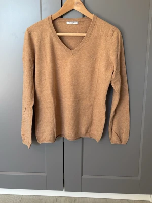 Beige V-ringad tröja Sand - Snygg beige tröja från Sand med V-ringning och långa ärmar. Tröjan är stickad i ett mjukt material och har diskret logga på bröstet. Perfekt för en stilren och avslappnad look.
