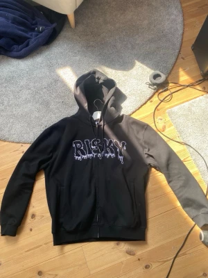 Svart hoodie med tryck 'RISKY' - Svart hoodie med dragkedja och huva, framtill finns ett stort vitt tryck med texten 'RISKY' i droppande stil. Mjuk bomullskänsla och ribbade muddar vid ärmslut och nederkant. Perfekt för en avslappnad streetwear-look.