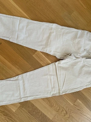 Beiga linnebyxor från Selected Homme - Snygga beiga linnebyxor från Selected Homme med avslappnad passform och resår i midjan. Byxorna har dragkedja och knappstängning samt diskreta fickor. Perfekta för en chill och stilren look under varma dagar.