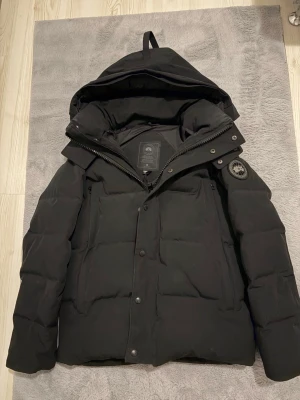 Svart dunjacka från Canada Goose - Säljer en svart dunjacka från Canada Goose med stor huva och patch på ärmen. Jackan har dragkedja, knappar och flera fickor. Perfekt för kalla vinterdagar och riktigt varm tack vare dunfyllningen.