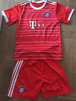 Bayern München Musiala matchställ stl: 146-152 - Rött Bayern München matchställ med Musiala #42. Tröjan har vita ränder, klubbmärke och sponsorlogga framtill samt namn och nummer på ryggen. Korta ärmar och shorts med vita ränder på sidorna. Tillverkat i lätt och ventilerande material.