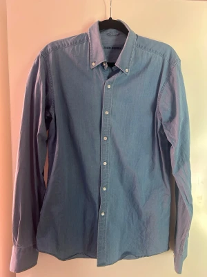 Blå skjorta med knappar från John Henric  - Klassisk blå skjorta i bomull med lång ärm och vita knappar hela vägen framtill. Skjortan har button-down krage och enkel, stilren design som passar till det mesta. Perfekt för dig som gillar en clean och tidlös look.