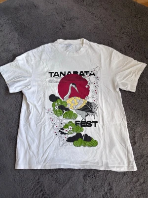 Tanabata  - Vit t-shirt med färgglatt japanskinspirerat tryck fram och bak, med texten 'Tanabata Fest', en trana, röd sol och gröna blad. Klassisk passform och rund halsringning. Perfekt för dig som gillar unika och grafiska motiv.