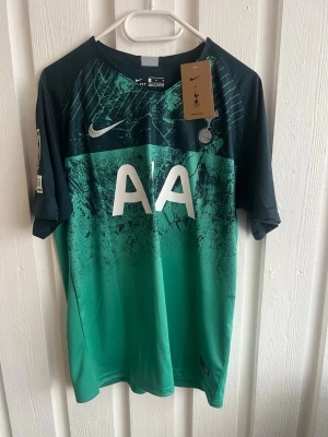 Tottenham bortatröja Lucas 27 Nike - Snygg Tottenham Hotspur bortatröja från Nike med grönt och svart mönster, tryckta detaljer som 'AA' på framsidan och 'Lucas 27' på ryggen. Champions League-märke på ärmen. Perfekt för fotbollsfans som vill sticka ut på planen eller läktaren.