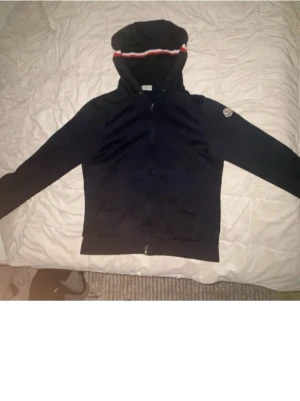 Svart hoodie från Moncler med dragkedja - Snygg svart hoodie från Moncler med hel dragkedja framtill och huva med rödvit rand. Moncler-logga på ärmen ger en exklusiv touch. Tillverkad i mjukt material som känns skönt mot huden. Perfekt för dig som vill ha en stilren och trendig hoodie.