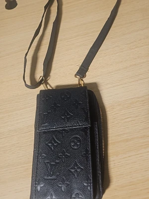 Svart axelväska  - Snygg svart axelväska från  i präglat skinn med klassiskt monogrammönster. Väskan har justerbar axelrem, dragkedja och flera fack för smart förvaring. Perfekt för dig som vill ha en stilren och lyxig accessoar.