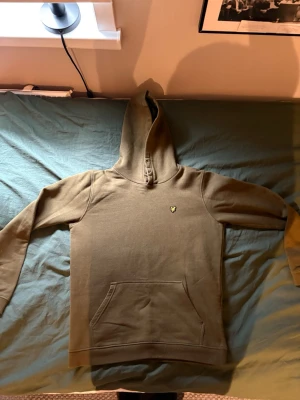 Olivgrön hoodie från Lyle & Scott - Olivgrön hoodie från Lyle & Scott med klassisk gul logga på bröstet. Hoodien har känguruficka framtill och ribbade muddar vid ärmslut och nederkant. Tillverkad i mjukt bomullsmaterial, perfekt för en avslappnad stil. Junior i storlek M