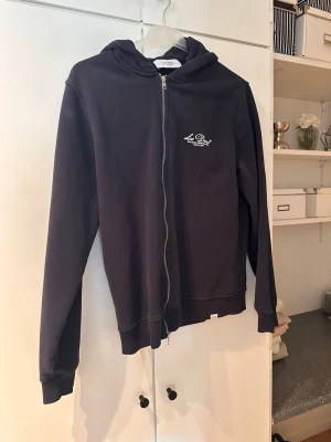 Mörkblå zip-hoodie Les Deux - Mörkblå hoodie med dragkedja från Les Deux. Klassisk modell med huva och ribbade muddar. Liten vit logga tryckt på bröstet. Perfekt för en chill och stilren look. Tillverkad i mjukt bomullsmaterial.