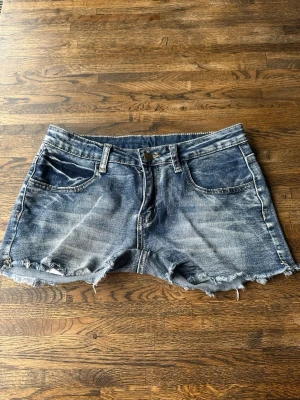 Blå jeansshorts med rå kant - Snygga blå jeansshorts med slitna detaljer och råa kanter. Shortsen har klassisk femficksdesign, bälteshällor och broderade bakfickor med mönster. Perfekta för sommaren och ger en avslappnad vibe.