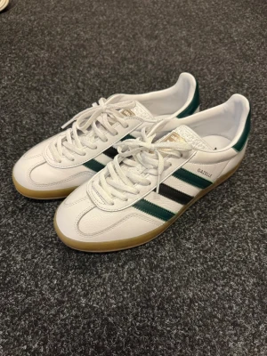 Adidas Gazelle vita sneakers - Säljer ett par klassiska Adidas Gazelle sneakers i vitt läder med gröna och svarta ränder på sidan. Endast använda 1 gång. Skorna har gummisula i ljusbrunt och vita skosnören. Diskret gulddetalj med texten 'GAZELLE' på sidan. Perfekt retrostil för dig som gillar streetwear. Saknas en liten hörna på flärpen av tungan men inget som varken syns eller känns.