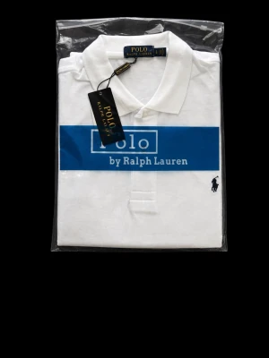 Vit pikétröja från Polo Ralph Lauren - Klassisk vit pikétröja från Polo Ralph Lauren med broderad logga på bröstet och krage med knappar. Tröjan har korta ärmar och är tillverkad i mjuk bomull. Perfekt för en stilren och avslappnad look.