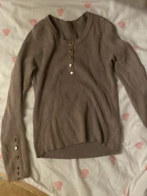 Beige ribbad långärmad tröja - Snygg beige ribbad tröja med lång ärm och rund halsringning. Tröjan har dekorativa knappar framtill och även på ärmsluten, vilket ger en trendig touch. Perfekt att matcha med jeans eller kjol för en stilren look.