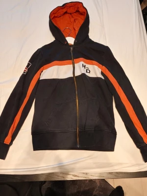 Harley-Davidson hoodie med dragkedja - Svart hoodie med orange och vita detaljer från Harley-Davidson. Dragkedja framtill, huva med orange insida och tryck på både bröstet och ryggen. Snygga orange ränder längs ärmarna och praktiska fickor framtill. Perfekt för dig som gillar MC-stil.