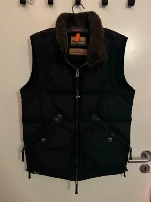 Svart dunväst Parajumpers med riktig päls - Stilren svart dunväst från Parajumpers med dragkedja framtill, två snedställda fickor med knappar och en hög krage med brun päls. Västen har detaljer i läder och märkets logga på nacken. Perfekt för lager-på-lager under kalla dagar. Kvitto tillkommer ej