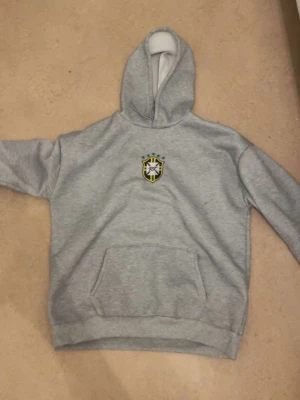 Grå hoodie med emblem - Snygg grå hoodie med huva och magficka. På bröstet finns ett broderat emblem i svart, vitt och gult med stjärna och sköld. Perfekt för en chill och avslappnad stil. Mjuk och skön bomullsblandning.