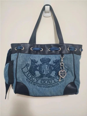 Juicy Couture daydreamer blå denim handväska - Snygg handväska från Juicy Couture i blå denim med svarta detaljer i konstläder. Väskan har broderad logga framtill, silverfärgade metalldetaljer och en dekorativ nyckelberlock. Praktiska handtag och sidoficka gör den både trendig och funktionell.