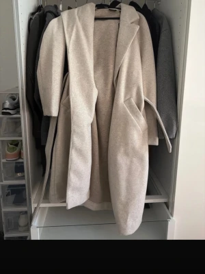 Beige lång kappa från Vero Moda - Stilren och lång beige kappa från Vero Moda med skärp i midjan och stora slag. Kappan har två fickor framtill och är tillverkad i ett mjukt material som ger en snygg och avslappnad look. Perfekt för dig som vill ha en klassisk och trendig ytterjacka.