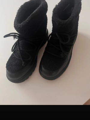 Svarta moon boots med teddyfoder - Säljer ett par svarta moon boots med mysigt teddyfoder och grov sula. Skorna har snörning framtill och en cool, chunky design som passar perfekt till vintern. Materialet är en mix av textil och syntet, vilket gör dem både varma och tåliga.