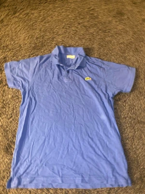 Blå pikétröja från Lacoste - Säljer en klassisk blå pikétröja från Lacoste med korta ärmar och den ikoniska krokodilloggan på bröstet. Tröjan har krage och knappar framtill. Tillverkad i mjuk bomull som känns skön mot huden.