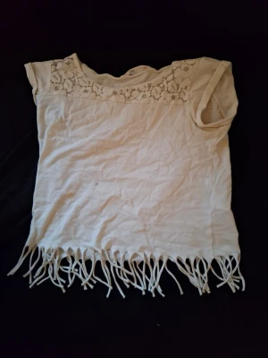 Vit topp med spets och fransar - Säljer en vit topp med korta ärmar, spetsdetaljer över axlarna och snygga fransar längst ner. Toppen har en avslappnad passform och är gjord i ett mjukt tyg, perfekt för en chill och trendig look. Från H&M. Har hål och en fläck