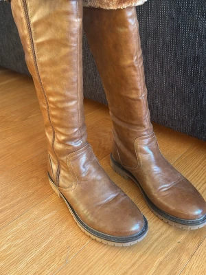 Bruna höga läderstövlar med dragkedja - Snygga bruna stövlar i konstläder med högt skaft och grov sula. Stövlarna har dragkedja på sidan och en klassisk, tidlös design som passar till många olika outfits. Perfekta för våren  när du vill ha både stil och komfort. Endast använda ett fåtal gånger. 