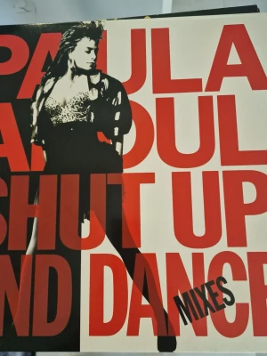 Vinyl: Shut Up and Dance Mixes - Vinylskiva med albumet 'Shut Up and Dance (Mixes)' av Paula Abdul. Omslaget har stora röda bokstäver och en svartvit bild på artisten. Baksidan listar låtar som Coldhearted, Straight Up och Opposites Attract. Perfekt för samlare av pop och dansmusik. Inga repor.