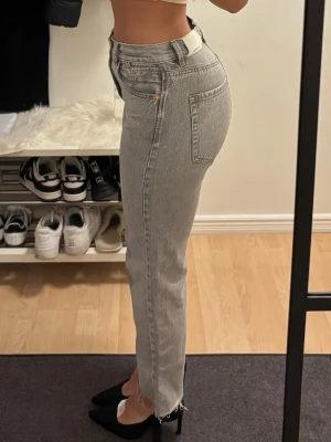 Ljusgrå raka jeans med rå kant - Säljer ett par ljusgrå raka jeans med hög midja och rå, fransad kant nedtill. Jeansen har klassiska fem fickor och stängs med knapp och dragkedja. Perfekta för en avslappnad och trendig look.