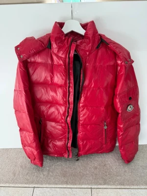 Röd pufferjacka  - Snygg röd pufferjacka från Moncler med avtagbar huva och två dragkedjefickor framtill. Jackan har quiltad design, hög krage och logotyp på ärmen. Tillverkad i glansigt material som ger en modern look. Perfekt för kalla dagar.