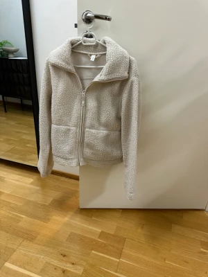 Beige teddyjacka med dragkedja - Mysig beige teddyjacka med hög krage och dragkedja framtill. Jackan har två stora fickor fram och är gjord i fluffigt, mjukt material som ger en avslappnad och trendig look. Perfekt för kyliga dagar när du vill vara både varm och snygg.