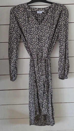 Blommig klänning från Saint Tropez - Snygg blommig klänning från Saint Tropez i svart och vitt. Klänningen har v-ringning, lång ärm och ett knytband i midjan för en smickrande passform. Perfekt för dig som gillar feminina detaljer och mönster.