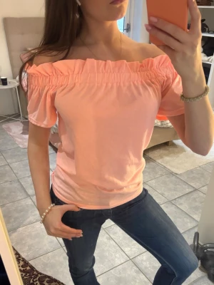 Corelli offshoulder topp - Trendig ljusrosa/corall offshoulder topp med korta ärmar och smockad kant upptill. Perfekt för dig som vill ha en söt och enkel topp till jeans eller kjol. Tillverkad i mjuk bomull som känns skön mot huden.